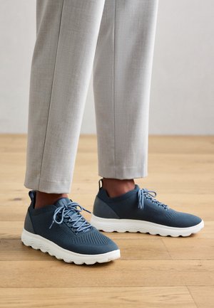 Sneakers blu a maglia con una tomaia testurizzata, chiusura con lacci e suola in gomma bianca. Indossati con pantaloni grigi chiari su un pavimento in legno.