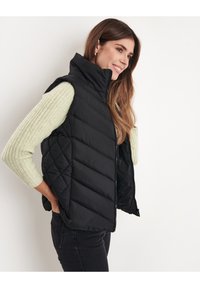 Gilet puffo nero con alto collo, design trapuntato e chiusura con zip, indossato sopra un maglione a coste verde chiaro.