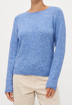 Vrouw draagt een lichtblauwe, gebreide trui met lange mouwen gecombineerd met beige broek, getoond van nek tot heupen tegen een effen achtergrond.