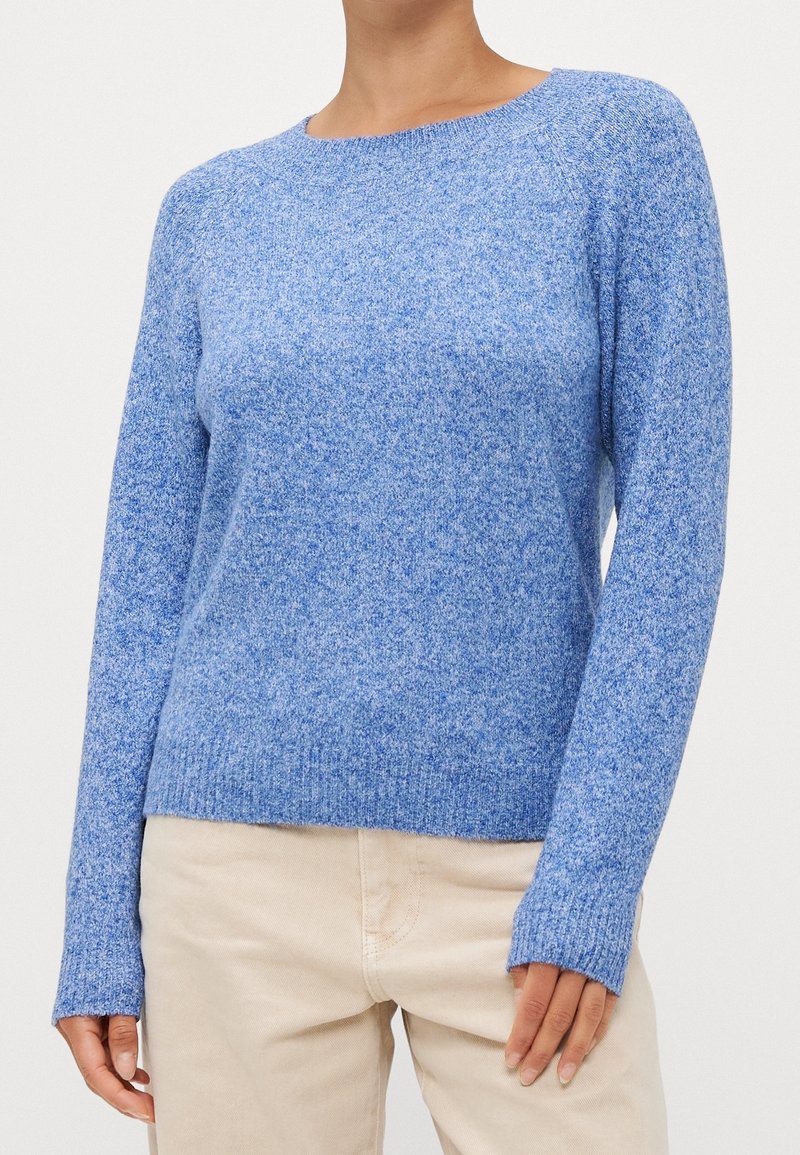 Femme portant un pull en maille bleu clair à manches longues associé à un pantalon beige, montrée du cou aux hanches sur un fond uni.