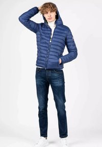 Giacca piumino blu con cappuccio e cuciture orizzontali. Abbinata a jeans scuri e sneakers bianche, il modello mostra un outfit casual.