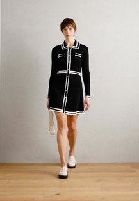 Robe noire en maille avec des bordures blanches, design collier, fermeture à boutons sur le devant, et longueur jusqu'aux genoux. Associée à des chaussures de couleur claire et un sac.