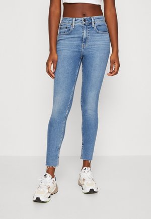 Personne portant un jean skinny taille haute bleu clair avec des ourlets effilochés et des baskets blanches beige, debout devant un fond blanc.