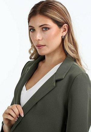 Olivgrüner Blazer mit geknöpftem Revers, getragen über einem weißen Oberteil. Glatte Textur, taillierter Schnitt, mit subtilen Schulterpolstern und Knopfdetails.