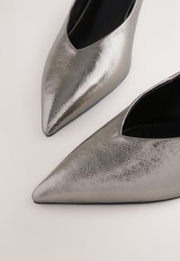 Ezüst színű, fémes, hegyes orrú balerinacipő pár fényes, texturált felülettel és fekete belső béléssel, világos háttéren.