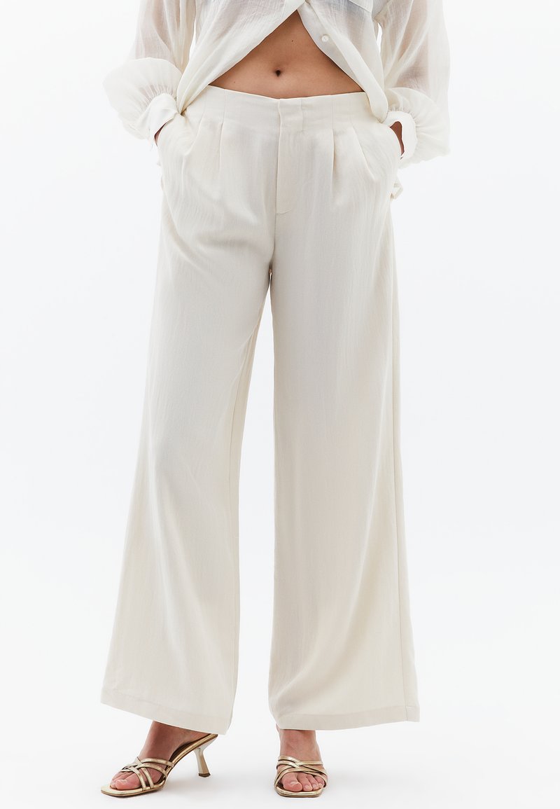 Breda byxor i ljus krämfärgad tyg med en mjuk textur, med framfickor och veck framtill. Stylad med guldiga sandaler med remmar.