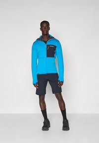 Kék cipzáras kapucnis pulóver texturált anyagból, fekete mellzsebbe dugva, fekete rövidnadrággal és túracipővel viselve. A modell egyenesen áll egy sima háttér előtt.