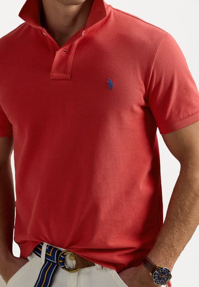 Sarkans polo krekls ar teksturētu audumu, ar zilā krāsā izšūtu logotipu uz kreisā krūšu. Valkāts ar baltām biksēm un svītrētu jostu.