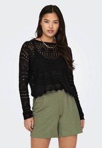 Svart åpenstrikket kort genser med bølget kant og intrikate mønstre, kombinert med olivengrønne shorts.