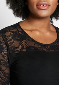 Haut noir avec des détails en dentelle sur les épaules et le décolleté. Tissu lisse avec une coupe ajustée, décolleté rond et motif floral en dentelle.