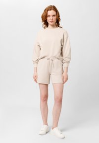 Béžová bavlněná mikina volného střihu s kulatým výstřihem, doplněná o odpovídající šortky s elastickým pasem. Celkový vzhled doplňují bílé tenisky.