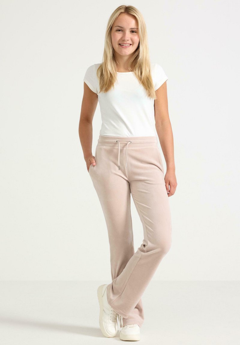 Jeune femme souriante, debout avec les mains dans les poches, portant un t-shirt blanc, un pantalon de survêtement beige et des baskets blanches sur fond uni.
