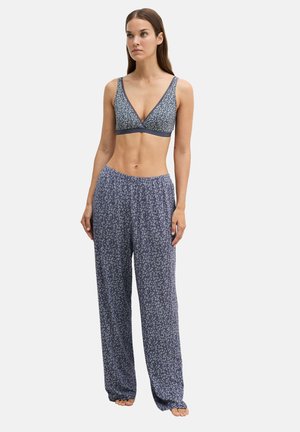 Donna in piedi scalza che indossa un bralette azzurro a fiori e pantaloni larghi azzurri a fiori abbinati su sfondo chiaro e uniforme.
