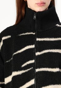 Kobieta w czarno-białej, paskowanej polarowej kurtce z wysokim prążkowanym kołnierzem i zapięciem na zamek błyskawiczny, odsłaniająca usta i częściowo szyję.