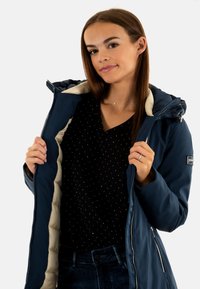 Giacca imbottita blu navy con interno crema, chiusura con zip e cappuccio. Abbinata a un top nero con piccole pois bianchi.