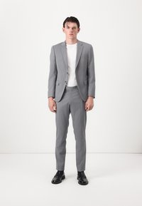 Costume gris avec des épaules structurées, deux boutons et revers en pointe, associé à un T-shirt blanc et des chaussures noires. Tissu lisse et coupe ajustée.
