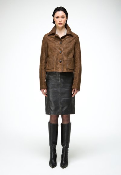 Femme debout, face à l'avant, portant une veste marron boutonnée, une jupe en cuir noire jusqu'aux genoux et des bottes noires montant jusqu'aux genoux.