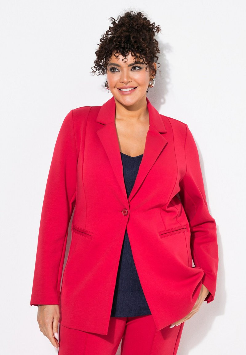 Rode blazer van zacht fabric, met een enkele knopen sluiting, reverskraag en twee voorzakken. Gecombineerd met een marineblauwe top.