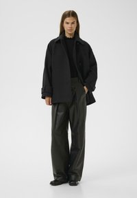 Dark wool coat with large collar and button closure, worn over a black sweater. Loose-fitting black leather pants and flat shoes.