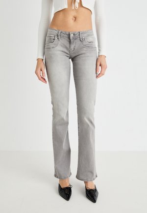 Jean gris clair slim-fit porté avec un top blanc court à manches longues et des chaussures noires pointues à lacets sur un fond uni.