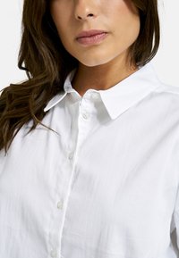 White button-up shirt with a soft texture, featuring a collared neckline and pearlescent buttons. No patterns or accents visible.