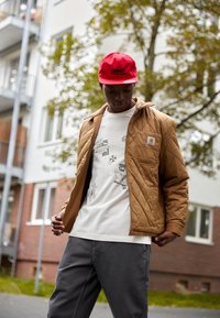 Chaqueta marrón acolchada con cierre tipo cremallera, llevada sobre una camiseta gráfica blanca. La persona lleva una gorra roja. En el fondo se puede ver un edificio de apartamentos.