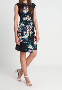 Schwarzes Blumenkleid mit tailliertem Design, das rosa und blaue Blumen zeigt, und kurzen Kappärmeln. Kombiniert mit weißen, schmalen Sandalen.