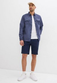 Camicia blu in chambray con chiusura a bottoni e due tasche sul petto, abbinata a pantaloni corti blu scuro e sneakers bianche. Il cappellino è di un beige chiaro.