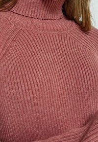 Pull en maille côtelée couleur mauve, avec un motif texturé et une silhouette ajustée, mettant en avant le col haut et les détails de couture.