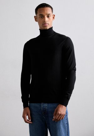 Maglione a collo alto nero in un design aderente, realizzato in un tessuto morbido e sottile a maglia, abbinato a jeans blu, mostrato su uno sfondo neutro.