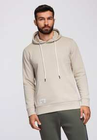 Béžová mikina s kapucí, přední kapsou, nastavovacími šňůrkami a černými akcenty. V kombinaci s tmavě zelenými tepláky pro neformální vzhled.