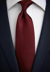 Bordeaux gekleurde, gestructureerde stropdas met een smalle vorm, geplaatst onder de kraag van een wit overhemd en naast een donkerblauw colbert.