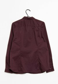 Chemise longue à manches maroon affichée sur un cintre noir, avec un col boutonné, des coutures cousues et un ourlet arrondi. Texture lisse.