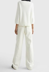 Person i lösa vita byxor och en vit topp med tre fjärdedels ärm, stående med ryggen mot betraktaren på en enfärgad bakgrund.