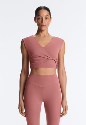 Femme portant un crop top portefeuille sans manches couleur rose et des leggings taille haute, se tenant face à l'avant avec une expression neutre.