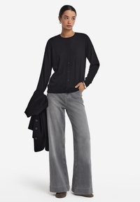 Svart cardigan med knappar, bärs över grå vida jeans. Slät textur, avslappnad passform, med långa ärmar. Grå stövlar och en svart tröja över axlarna.