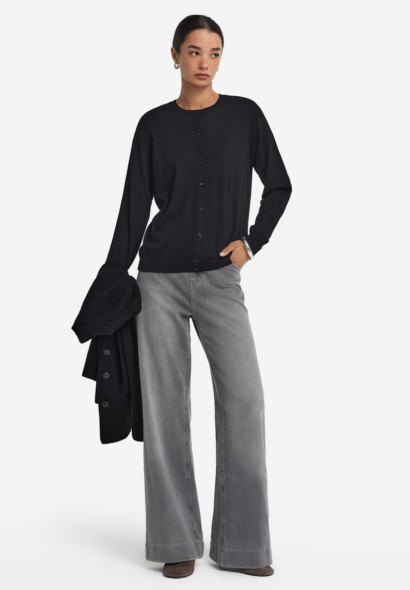 Svart cardigan med knappar, bärs över grå vida jeans. Slät textur, avslappnad passform, med långa ärmar. Grå stövlar och en svart tröja över axlarna.