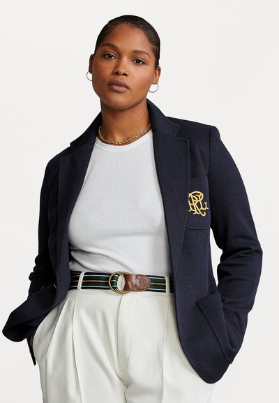 Blazer navy con un acabado texturizado, que presenta un logo bordado en oro en el bolsillo; combinado con una camisa blanca de canalé y pantalones crema.