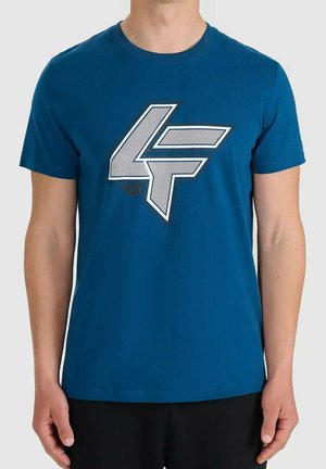 Mężczyzna ubrany w niebieski t-shirt z krótkim rękawem z dużym białym i czarnym obrysowanym nadrukiem "4F" na piersi, w połączeniu z czarnymi spodniami.