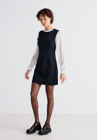 Musta hihaton mekko valkoisella pilkullisella pitkähihaisella paidalla. Mallilla on päällään mustat patenttijalkineet ja umpinaiset mustat sukkahousut. Minimalistinen muotoilu.