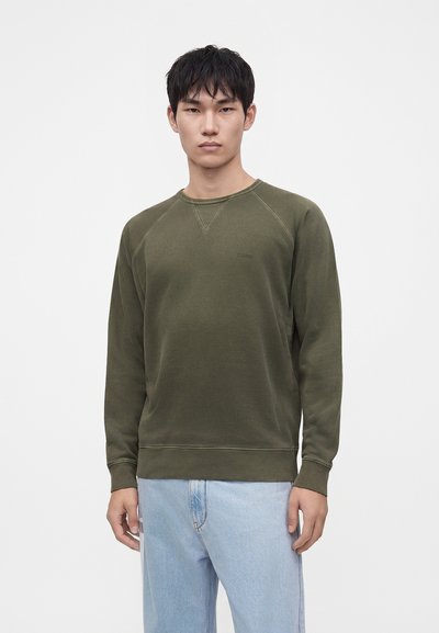 Jeune homme portant un sweat-shirt vert olive et un jean bleu clair, debout devant un fond clair uni.