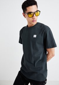 T-shirt in cotone verde scuro con scollatura rotonda, maniche corte e un piccolo etichetta sul petto. Modello che indossa occhiali da sole gialli.