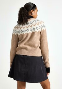 Brun strikket sweater med en geometrisk mønsterstrikning i creme og grå. Bæres over en sort flared nederdel, har lange ærmer og ribbede manchetter.