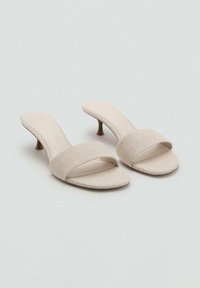 Mules à talons beiges en tissu avec une large bride avant et un design à bout ouvert, dotés d'un petit talon aiguille. Texture lisse et style minimaliste.