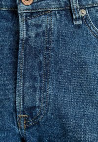 Jeans i middels blå vask med kontrasterende gul søm, med knappestang og synlige lomme-sømmer.