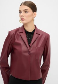 Blazer in ecopelle bordeaux con colletto a rever, chiusura a un bottone e taschino sul petto. Texture liscia e design corto.