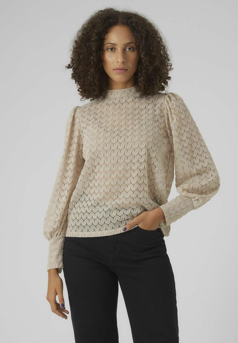 Blouse beige à motifs avec de longues manches bouffantes et un col montant. Tissage texturé avec un design géométrique, associée à un pantalon noir.