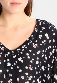 Schwarze Bluse mit V-Ausschnitt, aus leichtem Stoff, verziert mit einem Muster aus unterschiedlich großen weißen und rosa Punkten.