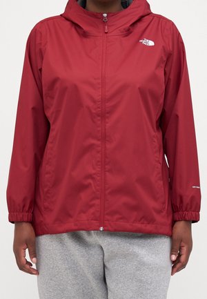 Czerwona kurtka przeciwdeszczowa na zamek błyskawiczny z kapturem, elastycznymi mankietami i logo The North Face na lewej piersi, noszona na szarych spodniach dresowych.