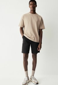 T-shirt in cotone beige e pantaloni corti neri, abbinati a sneaker beige e grigie. L'outfit presenta una vestibilità comoda, maniche corte e polsini a costine.